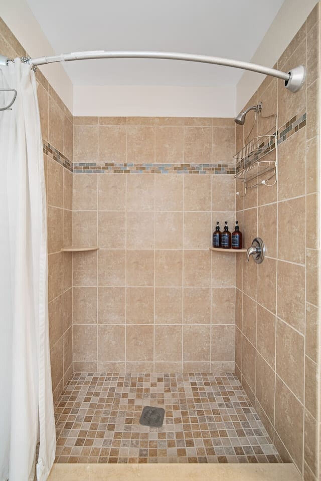Una ducha con azulejos color beige, una cortina de ducha blanca y un estante con artículos de aseo.