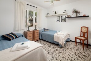 Un dormitorio con dos camas, un aparador de madera, una silla, un ventilador de techo y una ventana con cortinas.