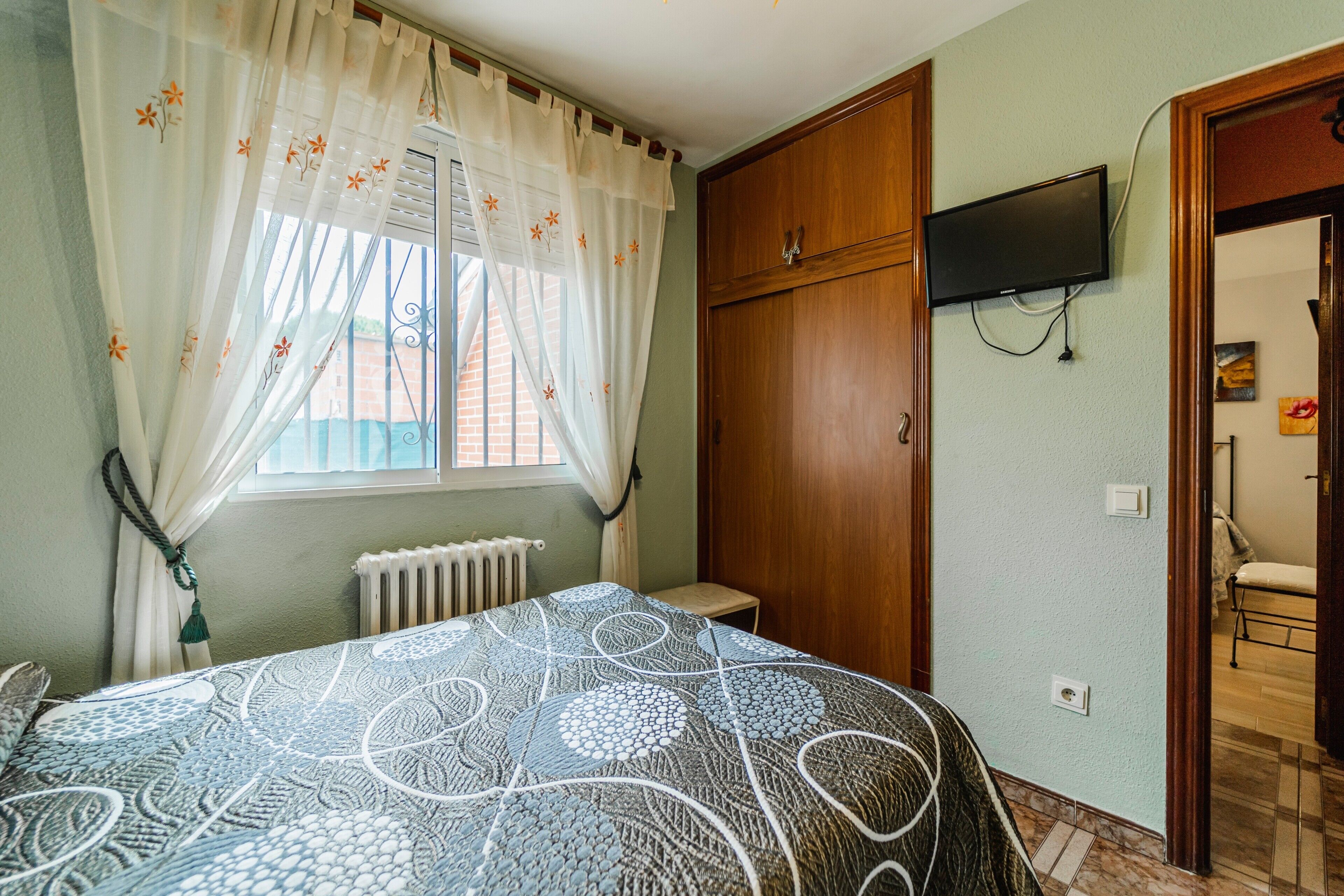 Une chambre à coucher comprenant un lit, un radiateur, une armoire en bois, un téléviseur fixé au mur et une fenêtre avec des rideaux.