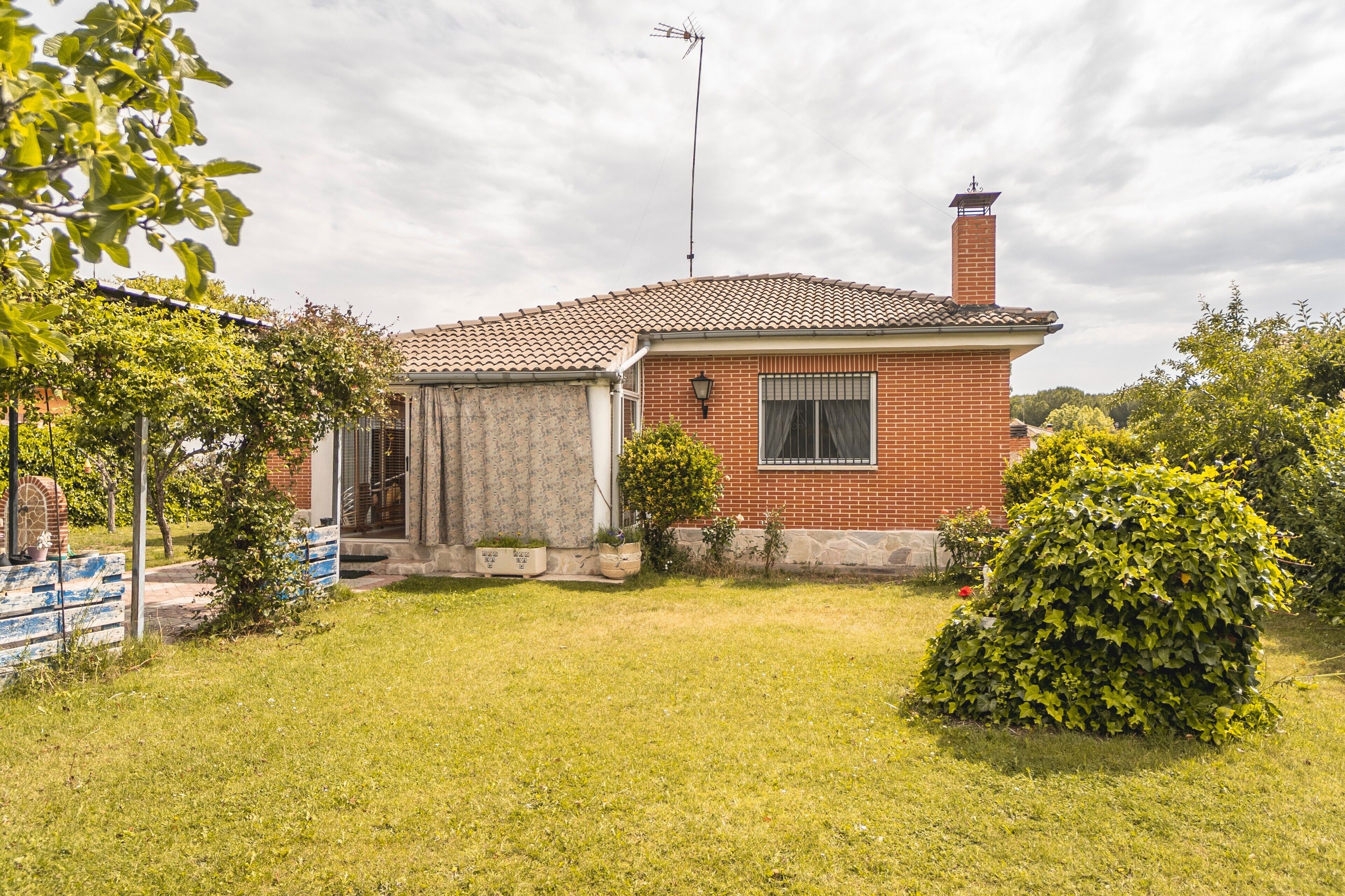 Une maison de plain-pied en briques, avec un toit en tuiles, une cheminée et un petit jardin devant, composé de pelouse et d’un arbuste.