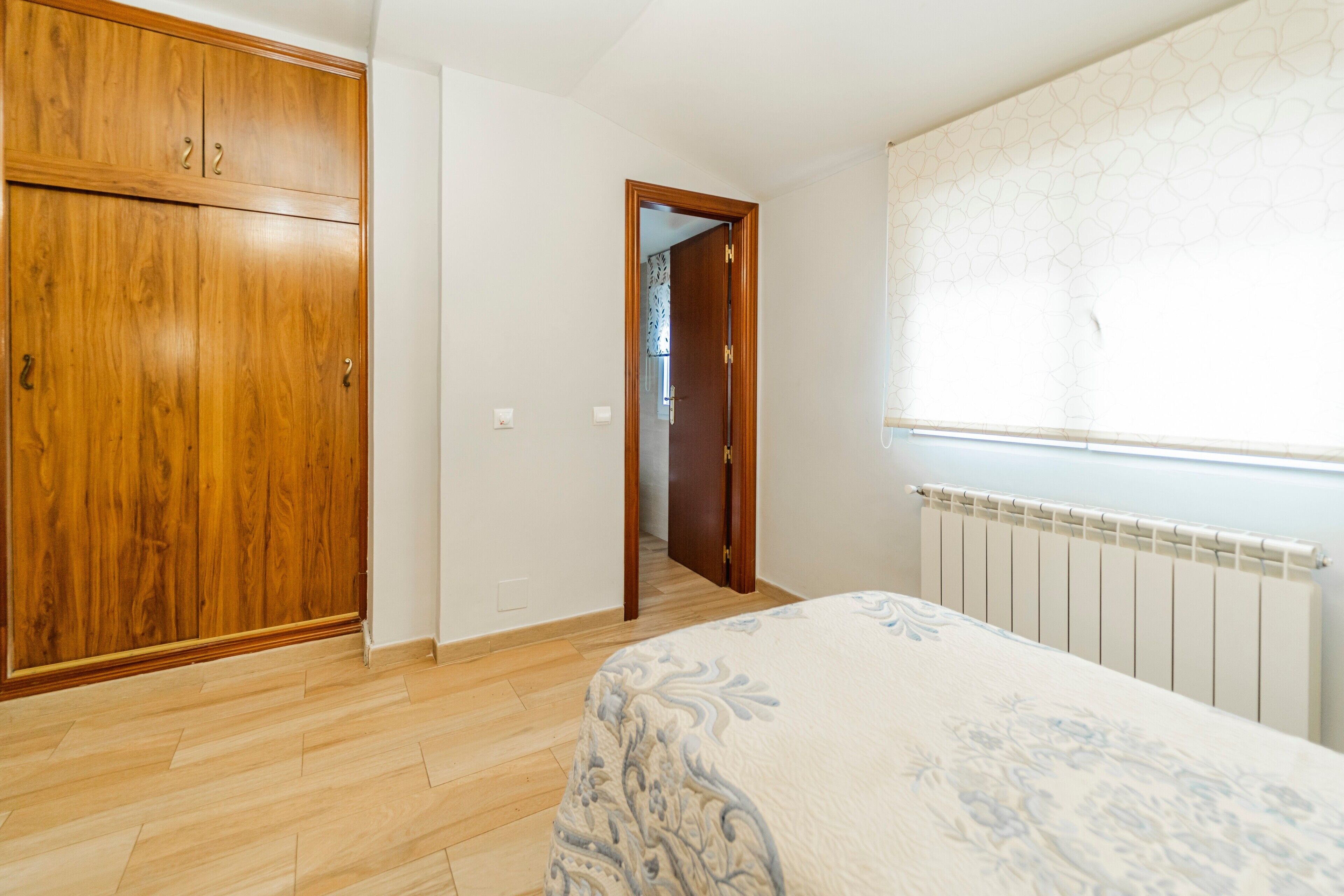 Un dormitorio con un armario de madera, una cama con colcha estampada, un radiador y una ventana con cortina decorada.