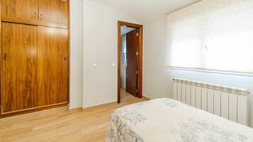 Un dormitorio con un armario de madera, una cama con colcha estampada, un radiador y una ventana con cortina decorada.