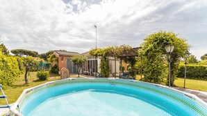 Une piscine dont le bord est revêtu de carrelage, entourée d’un jardin avec des arbres et un bâtiment en briques en arrière-plan.