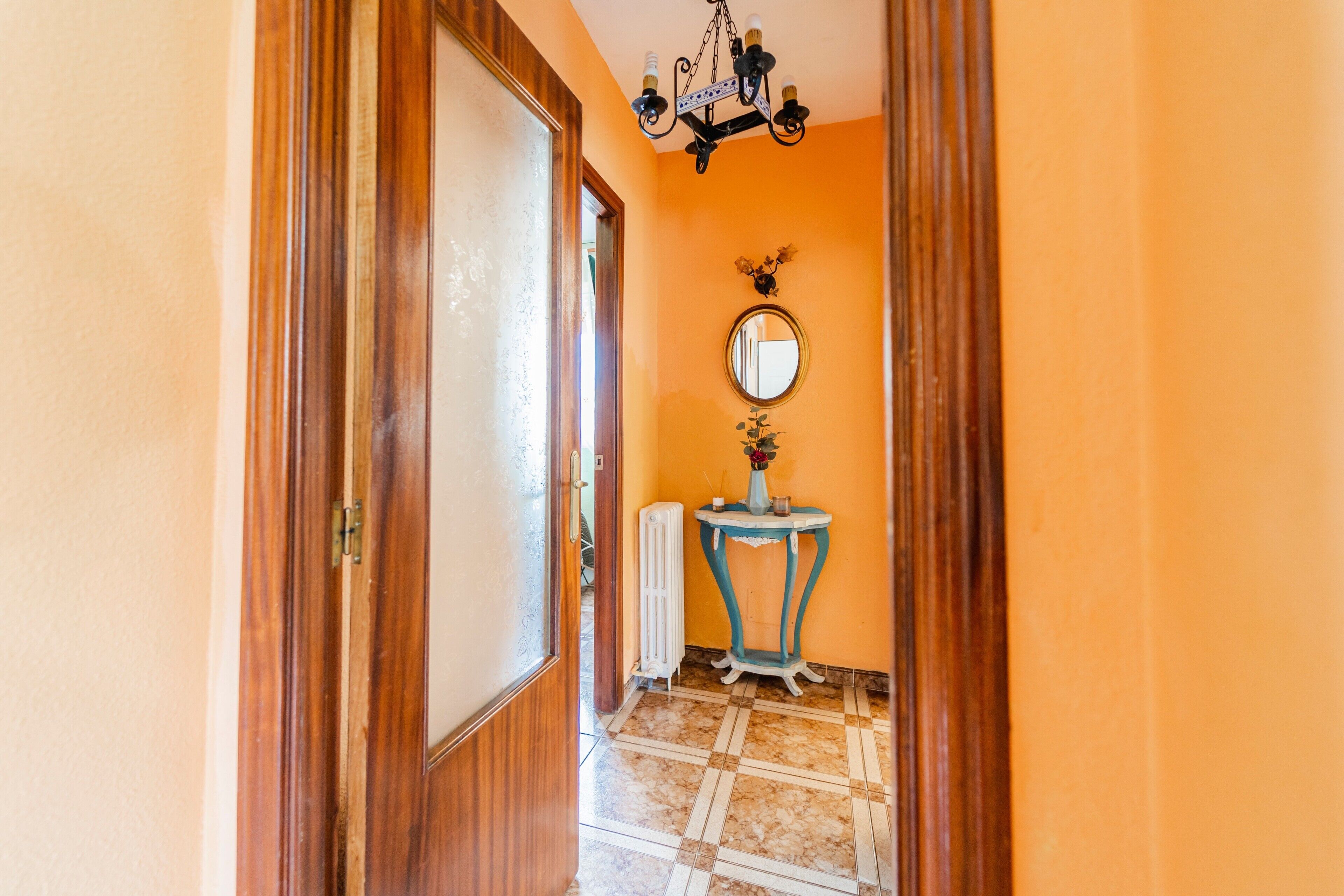 Un pasillo con puerta de madera, un radiador, una mesita con un florero y un espejo en la pared.