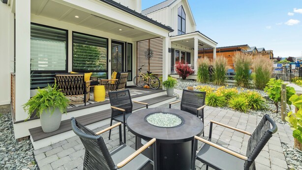 Een modern terras buiten met een ronde tafel en stoelen, omringd door planten in potten en een pad van stenen.