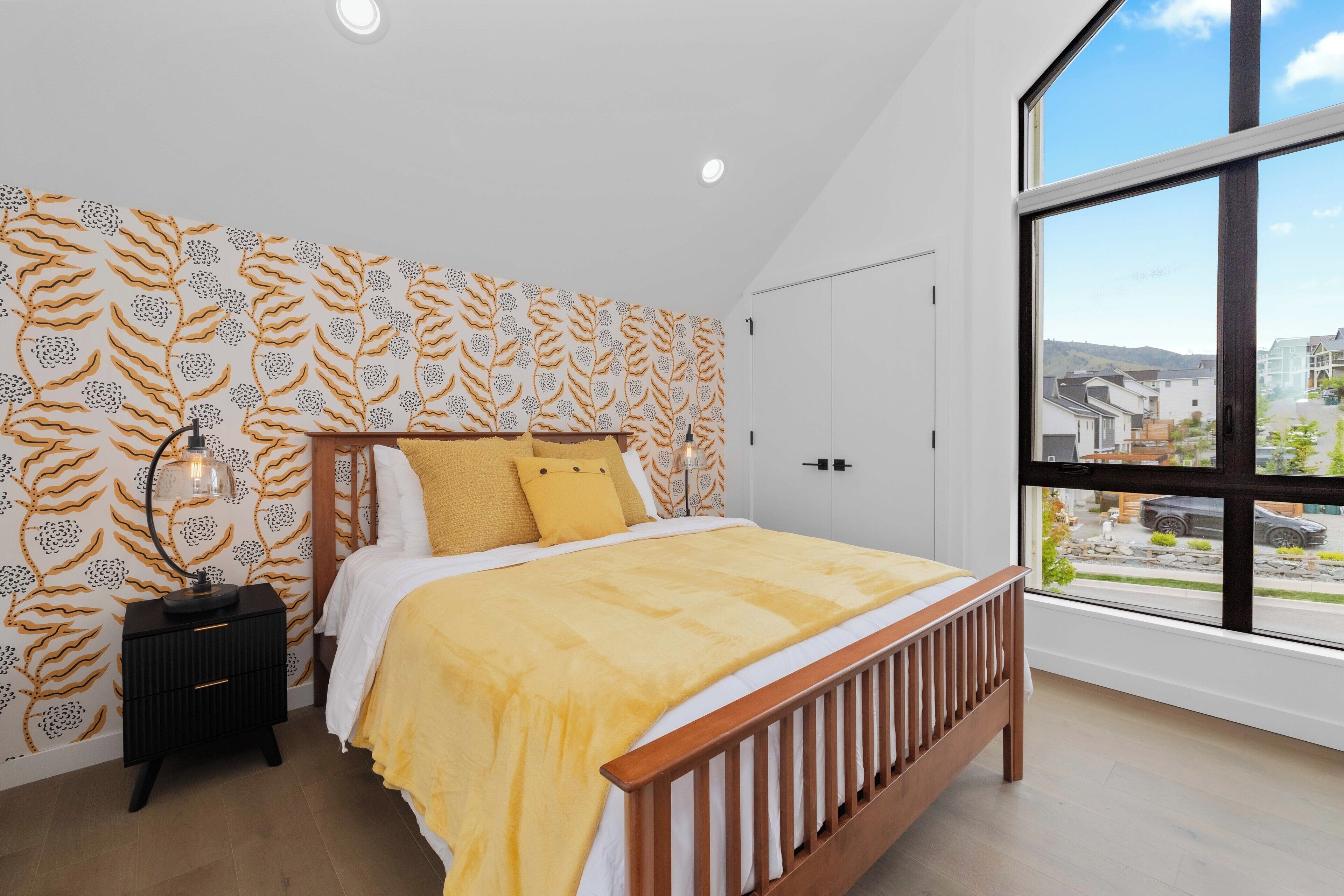 A bedroom with a large window, a bed with a wooden headboard, and patterned wallpaper.