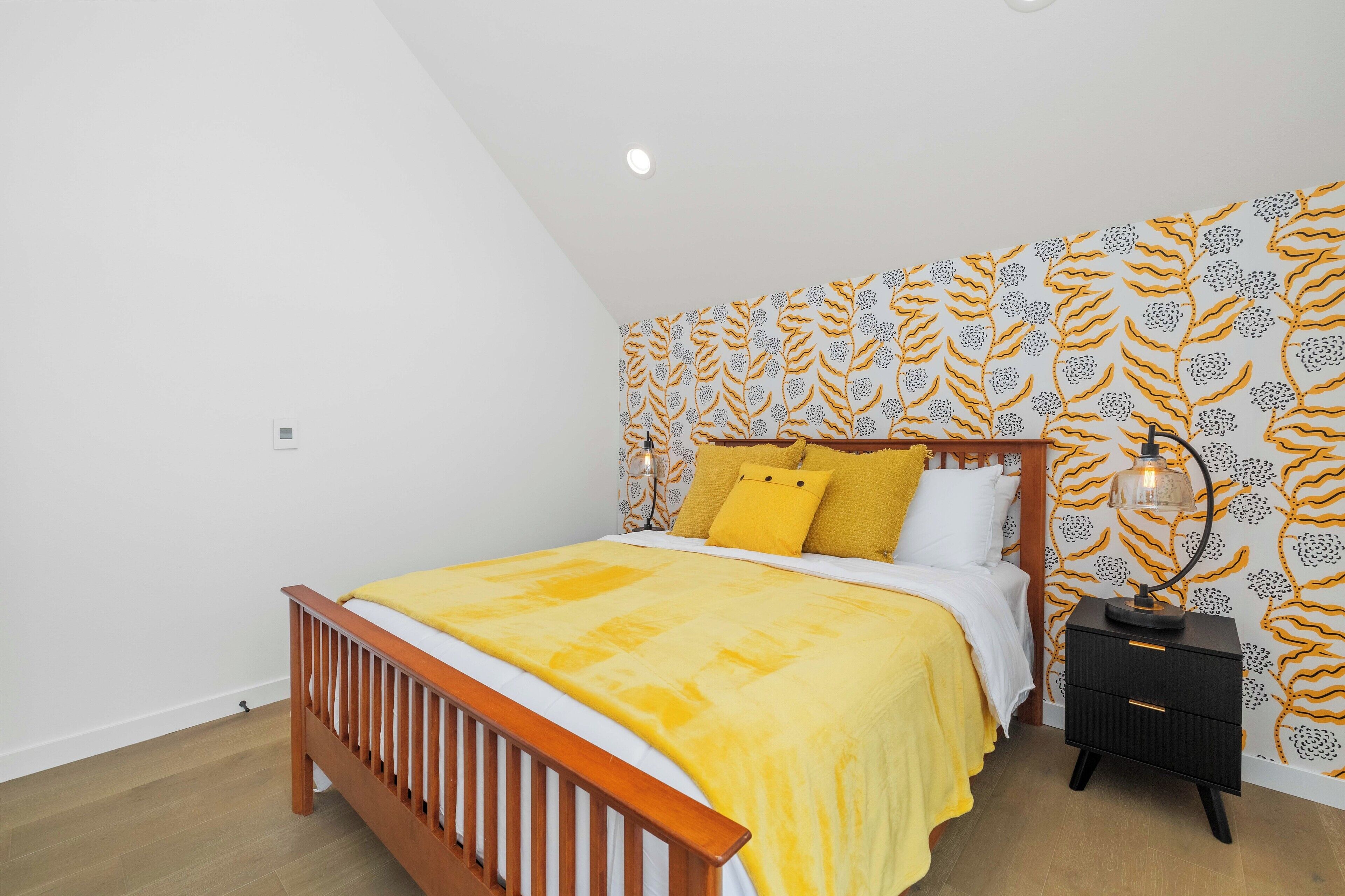 Une chambre à coucher avec un lit en bois, une literie jaune, une table de chevet avec une lampe et un papier peint à motifs.