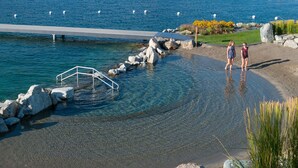 Een waterkant met een helder waterpad, een metalen trap die in het water leidt en een zandstrand met een grasveld en gele bloemen.