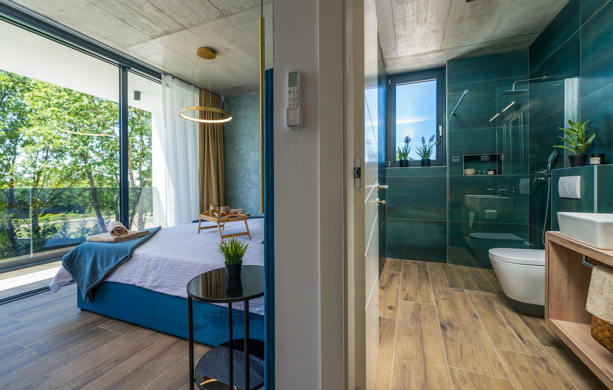 A modern bedroom with a large window, a bedside table, and a view of greenery. The bathroom features a glass shower enclosure and a wooden vanity.