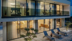 A modern two-story house with large glass windows, a balcony with lounge chairs, and a well-lit interior.