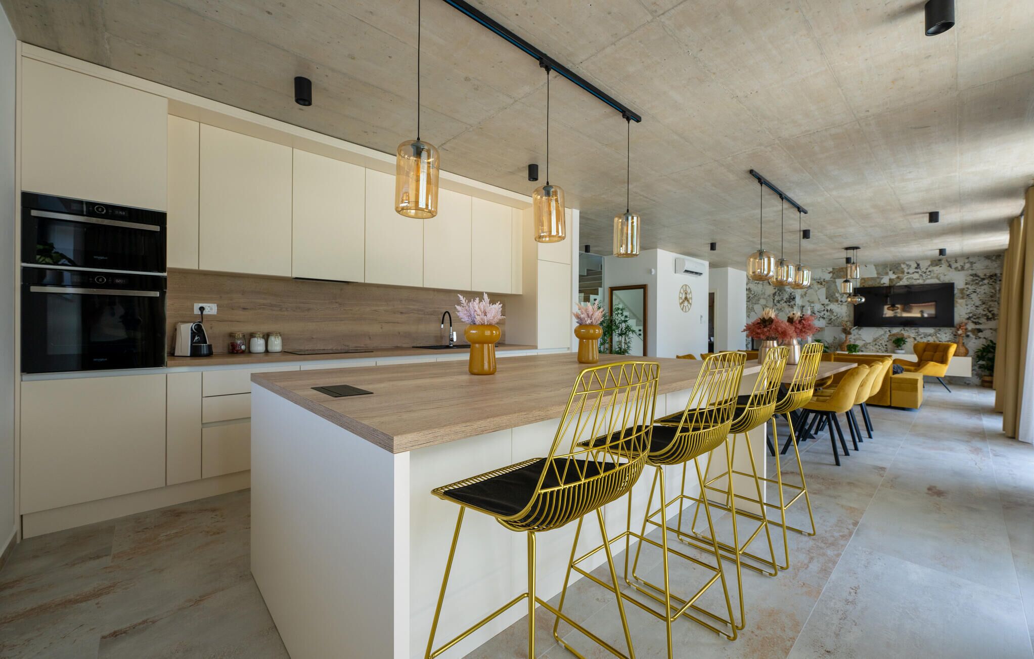 A modern kitchen with a large island, gold bar stools, pendant lighting, and a dining area with yellow chairs.