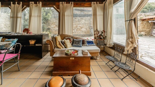 A cozy room with a wooden ceiling, a sofa with cushions, a coffee table, a dining table with chairs, and a view of a stone wall and greenery outside.