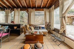 A cozy room with a wooden ceiling, a sofa with cushions, a coffee table, a dining table with chairs, and a view of a stone wall and greenery outside.
