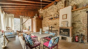 A rustic interior with a stone wall, wooden beams, a fireplace, and a dining area with chairs and a table.