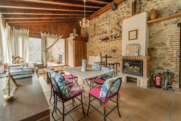 A rustic interior with a stone wall, wooden beams, a fireplace, and a dining area with chairs and a table.
