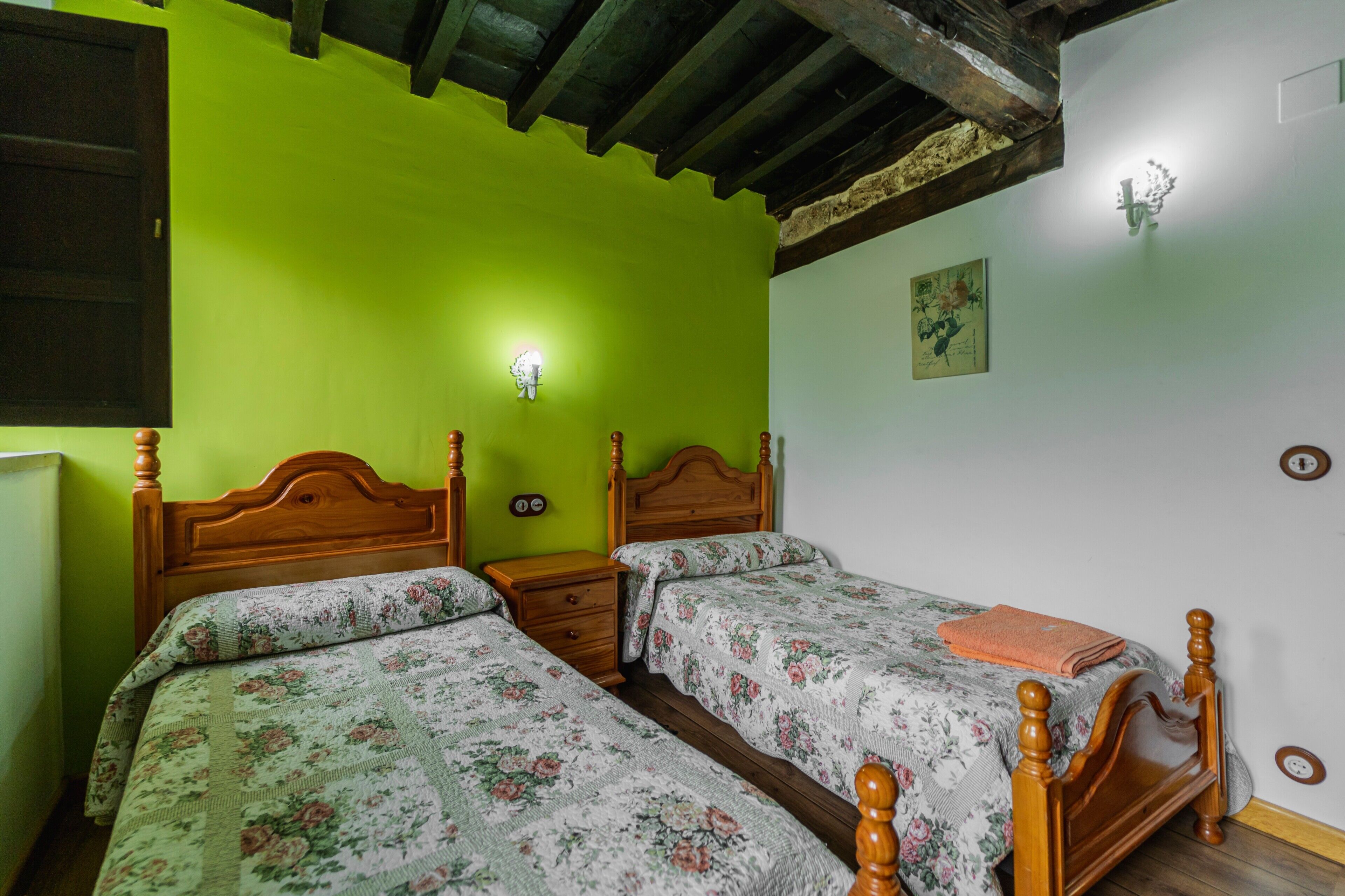 Quarto com duas camas de madeira, uma mesa de cabeceira, uma luminária na parede e um pequeno quadro.