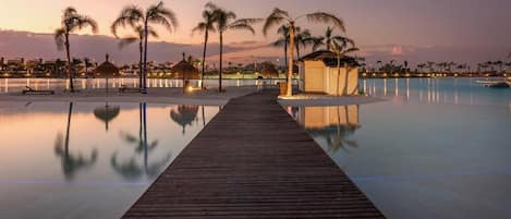 Una passerella di legno conduce a una piscina con palme e una piccola capanna, immersa in un’acqua tranquilla con uno skyline sullo sfondo.