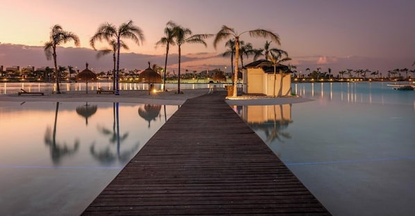 Una pasarela de madera conduce a una piscina con palmeras y una pequeña cabaña, rodeada por una masa de agua tranquila y un paisaje urbano al fondo.