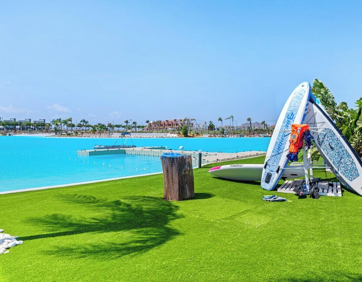 A poolside area with artificial grass, a wooden stump, and a surfboard leaning against a structure.