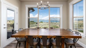 A dining room with a large wooden table and metal chairs, large windows with a view of a coastal landscape, and a chandelier. - Private Beach Access! Tennis/Pickleball Courts Nearby! Game Room! (Fish Haven)
