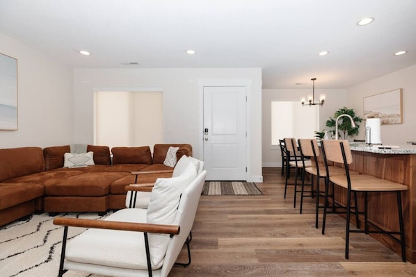 A living room with a brown sectional sofa, white armchairs, and a dining area with wooden chairs and a kitchen counter.