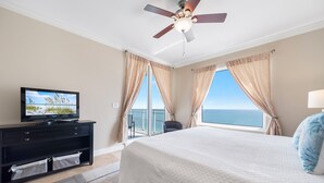 Un dormitorio con cama, televisor sobre una cómoda, ventilador de techo y vista al océano a través de amplios ventanales.