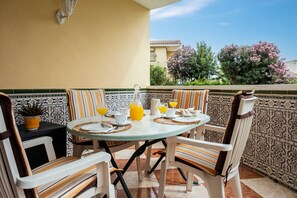 Ein runder Tisch mit Stühlen, eine Kanne Orangensaft und ein Glas Orangensaft auf einem Balkon mit gemusterten Fliesen und Blick auf Bäume und Gebäude.