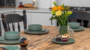 Een houten eettafel met groene borden en kommen, een glazen vaas met bloemen en twee cupcakes.
