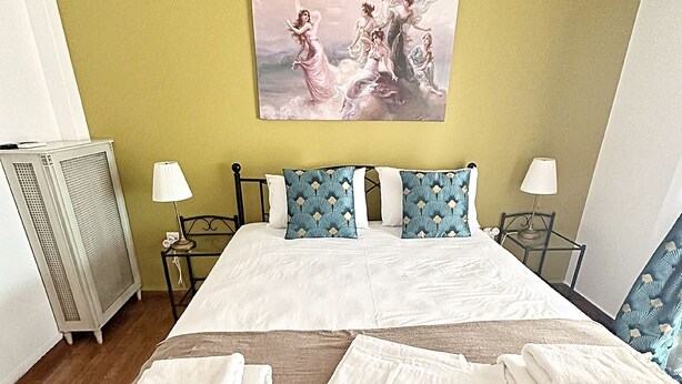 Cama bem arrumada com lençóis brancos e almofadas estampadas em azul, um quadro acima da cama e cabeceira com estrutura de metal.