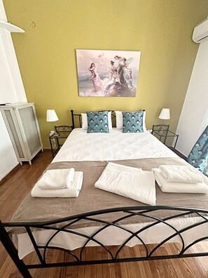 Cama bem arrumada com lençóis brancos e almofadas estampadas em azul, um quadro acima da cama e cabeceira com estrutura de metal.