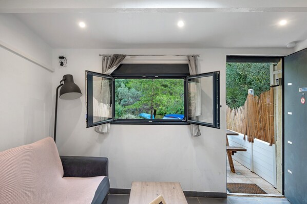 A room with a sofa, a table, a window with an open shutter, and a view of greenery outside.
