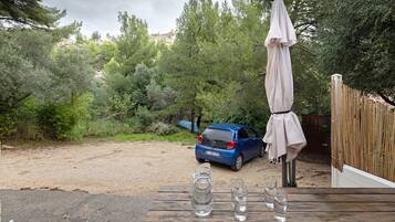 Un'auto parcheggiata su un’area sterrata con un pontile in legno, un tavolo con bicchieri e una brocca, e un ombrellone.