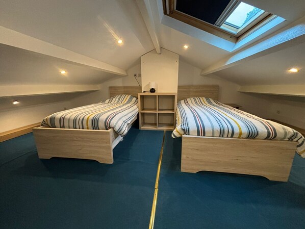 Une petite chambre mansardée, bien éclairée, avec deux lits en bois, une lucarne et une étagère centrale.
