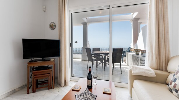 Ein Wohnzimmer mit Flachbildfernseher, Couchtisch aus Holz, einer Weinflasche und einem Glas. Eine Glastür verschiebt sich zu einem Balkon mit Tisch und Stühlen.