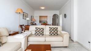 Ein Wohnzimmer mit einer Couch, zwei gemusterten Kissen, einem Holztisch, einer Lampe und einer Küche im Hintergrund.