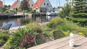 A waterfront patio with a wooden table and chairs, surrounded by greenery and a marina with boats.