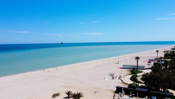 Ein Strand mit blauem Himmel, sandigem Ufer und einigen Gebäuden im Hintergrund.