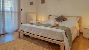 Een net opgemaakt bed met kussens en handdoeken, een nachtkastje, een wandlamp en een raam met dunne gordijnen.