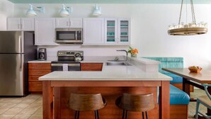 Una cocina moderna con armarios blancos, un refrigerador de acero inoxidable, un microondas y una cafetera. Hay una isla de madera con dos taburetes y una banca acolchada azul en la mesa de comedor.