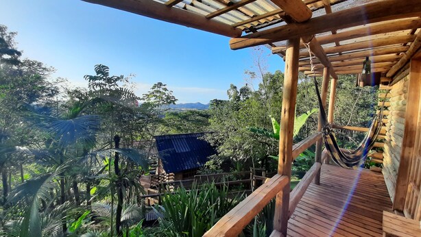 A wooden deck with a hammock, surrounded by lush greenery and a view of distant hills. - Chalé Tucano Eco.hospedagem - Virado Para o Nascer do Sol, com uma Vista Linda (Praia Grande)