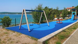 Une aire de jeux avec des balançoires et un toboggan, située près d’une étendue d’eau et d’un bâtiment.