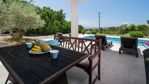 Une terrasse extérieure avec une piscine, des meubles en bois et une table garnie de fruits et de gobelets.
