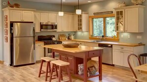 A kitchen with white cabinets, a stainless steel refrigerator, a wooden island with stools, a stove, and a window with a view of trees.