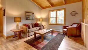 A cozy living room with a wooden ceiling, a sofa, a coffee table, a side table, a chair, and a window with a view.