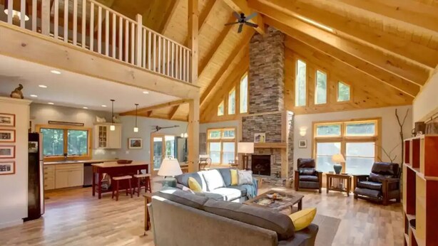 A spacious living area with a high ceiling, wooden beams, a stone fireplace, and large windows.