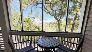 Une terrasse couverte avec une table et deux chaises, une poubelle et une vue sur des arbres et un bâtiment situé au loin.