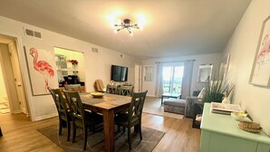 Un coin repas avec une table et des chaises en bois, un téléviseur en arrière-plan et un espace cuisine comprenant un réfrigérateur et un plan de travail.