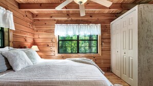 Un dormitorio de cabaña de madera con una cama, un ventilador de techo, una ventana con vistas a la vegetación y un armario.