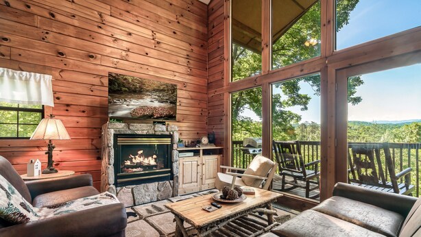 Interior acogedor de una cabaña con chimenea de piedra, muebles de madera y ventanas amplias que ofrecen vistas al exterior.