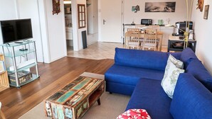 Un salon avec un canapé bleu, une table basse et un téléviseur posé sur un meuble. On y trouve également un coin repas avec une table et des chaises, ainsi qu’une cuisine équipée d’un four à micro-ondes et d’une machine à café.